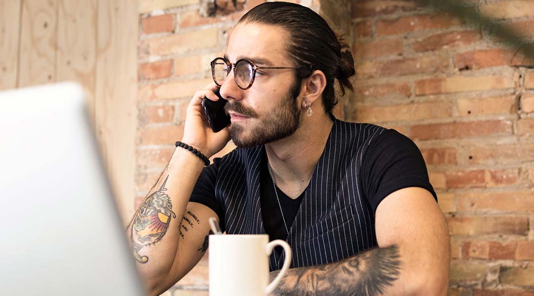 Portrait of bearded stylish man with tattooed arms in glasses making a phone call via cell sitting in coffee shop with cup of coffee and laptop on table